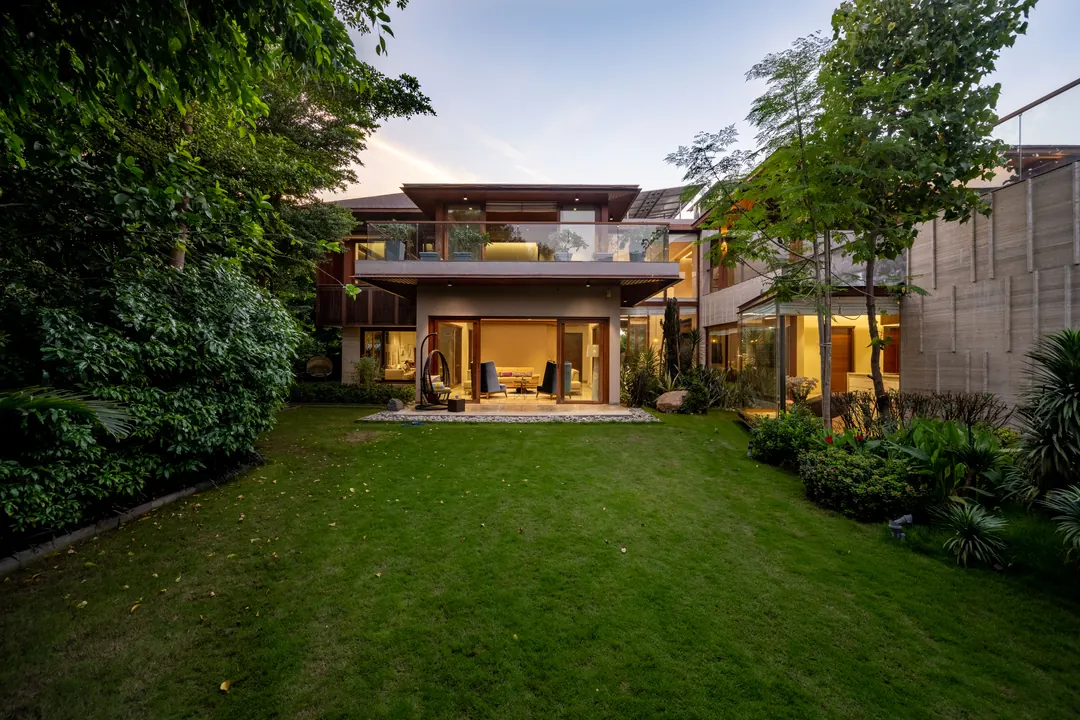 A timber house across the lawn at dusk, every room lit.