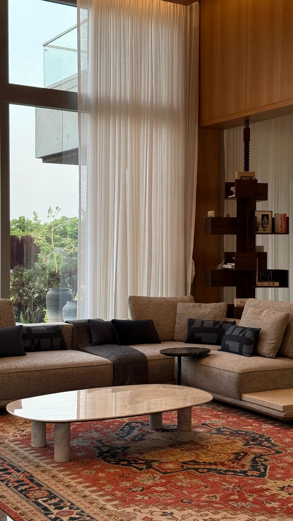 A double-height living room: tall curtains to the garden, a long sofa, a timber shelf.